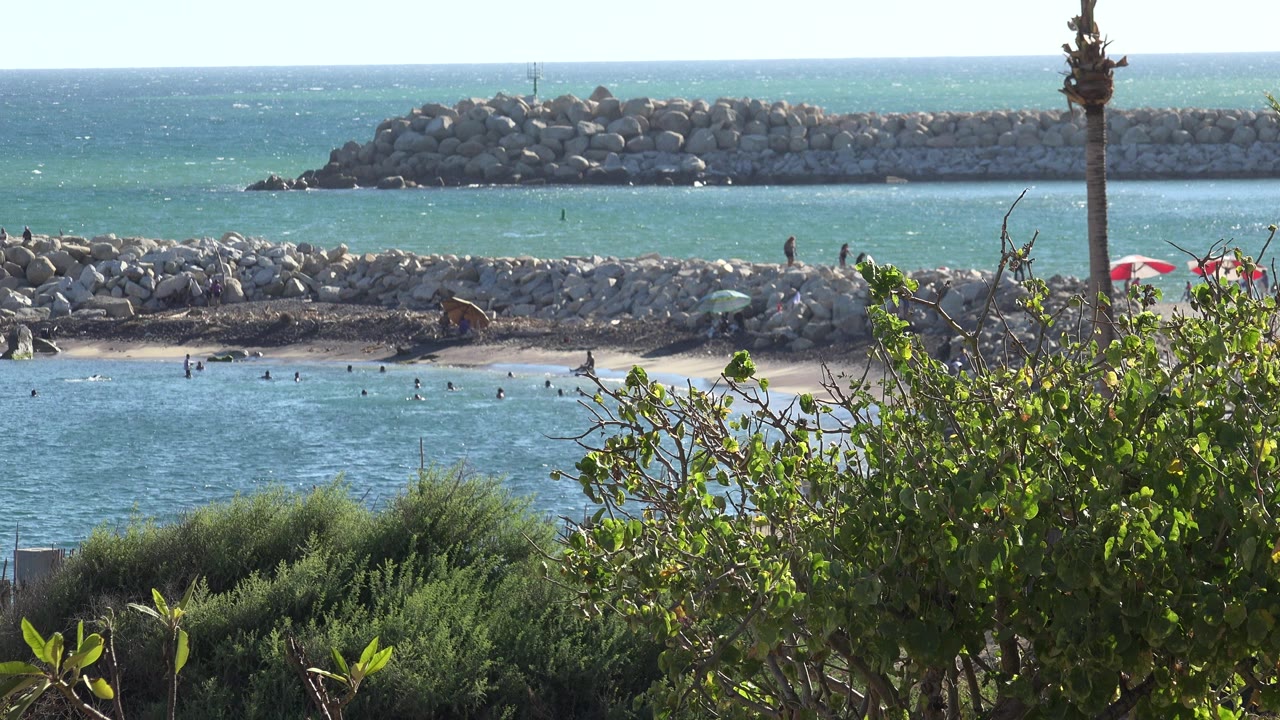 Opening frame of a Grand Junction, Colorado shoreline view with blue-green water, rock breakwaters, sand, shrubs in the foreground, and no identifiable people visible.