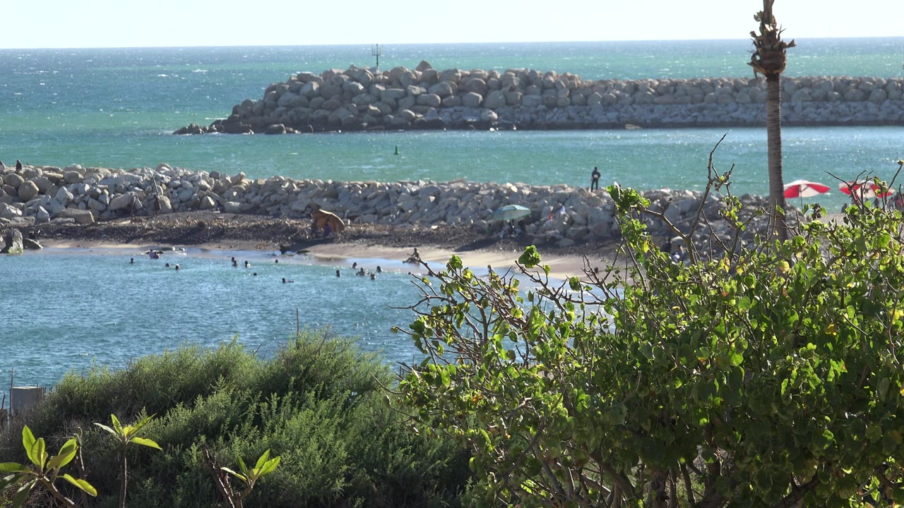 Grand Junction, Colorado shoreline one second in, showing choppy blue water, a sandy swimming area, small distant people, rock breakwaters, and leafy shrubs in front.