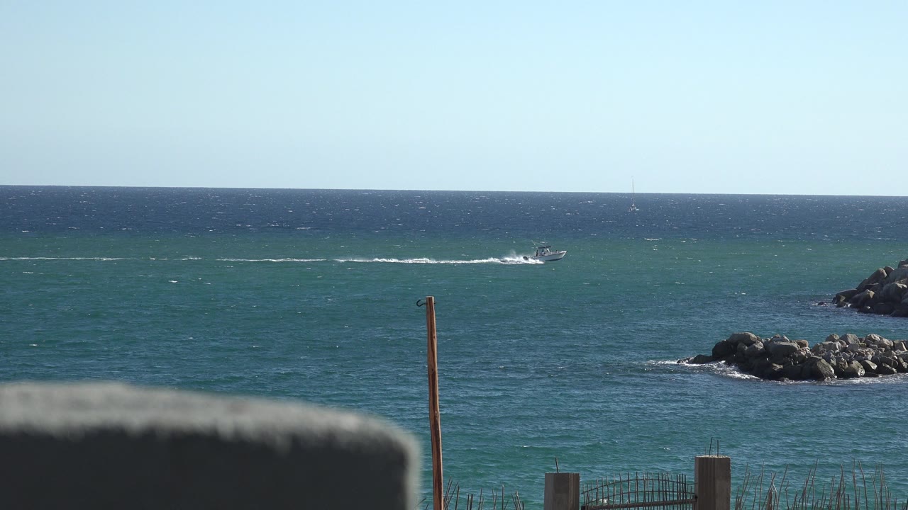Small boat on blue ocean beyond shoreline rocks and fence posts.