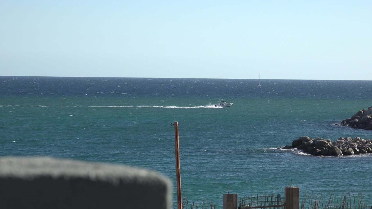 At 0:01, the boat sits farther along the distant waterline, leaving a white wake across the blue ocean while shoreline rocks and low fencing remain fixed in the foreground, with no visible people.