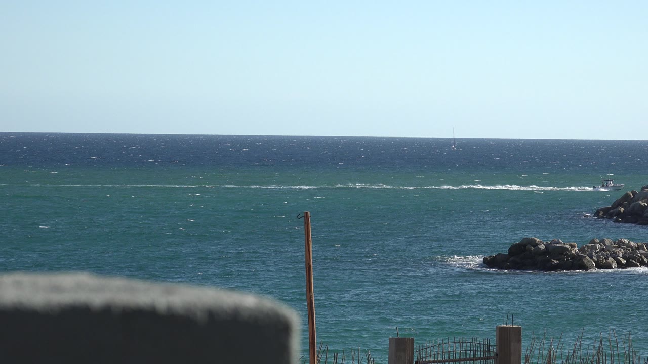 Opening frame from Grand Junction, Colorado footage showing blue sea, a rocky breakwater at right, thin posts near shore, a blurred dark foreground edge, and no visible people.