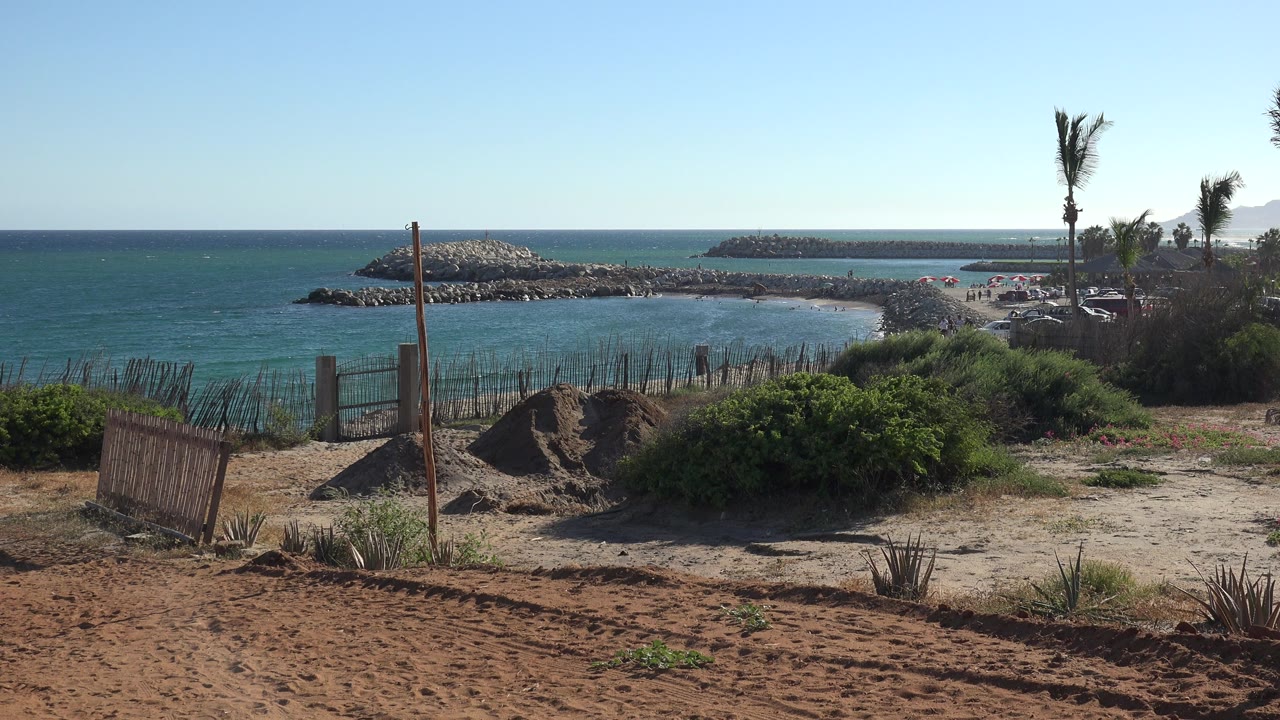 Sandy overlook with beach, blue water, rocky breakwaters, palms, and fencing.