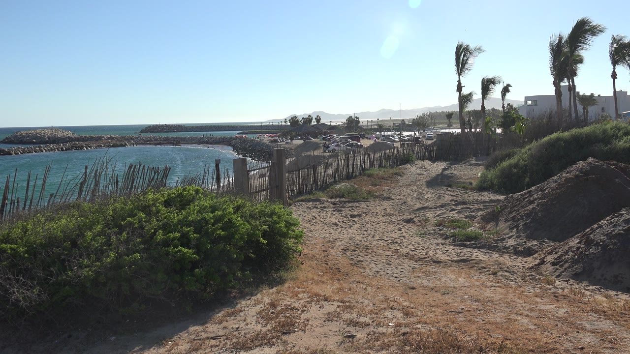 Opening frame of a Grand Junction, Colorado shoreline view with blue water at left, low shrubs and sandy ground in front, fencing and buildings beyond, and no visible people.