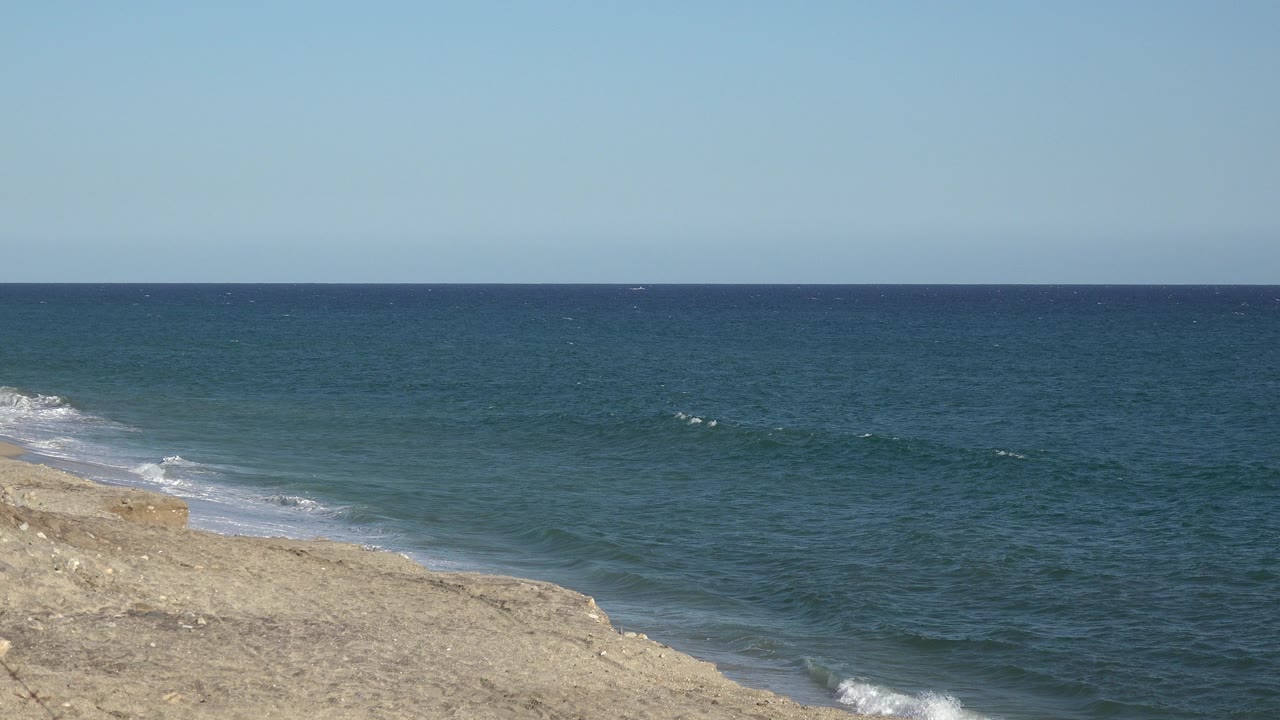 Blue ocean beside a tan shoreline under clear sky.