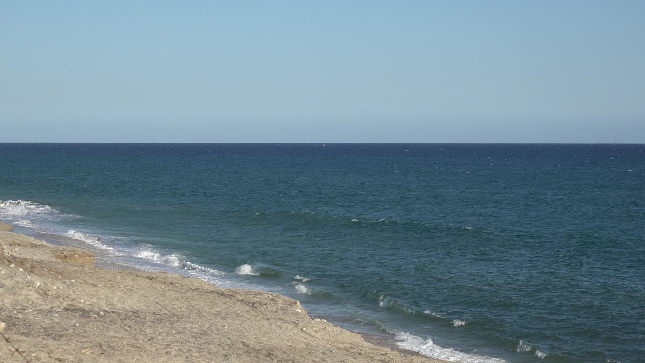 A brighter patch of white surf breaks near the tan shoreline while blue water stretches to the horizon in the Grand Junction Colorado clip, with no visible people.