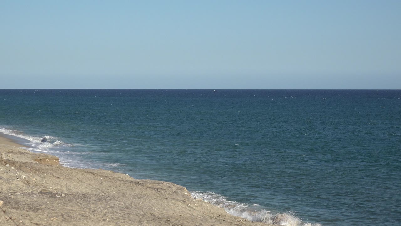 Opening frame of blue ocean water beside a tan beach or low shore under a pale clear sky in Grand Junction Colorado, with no visible people.