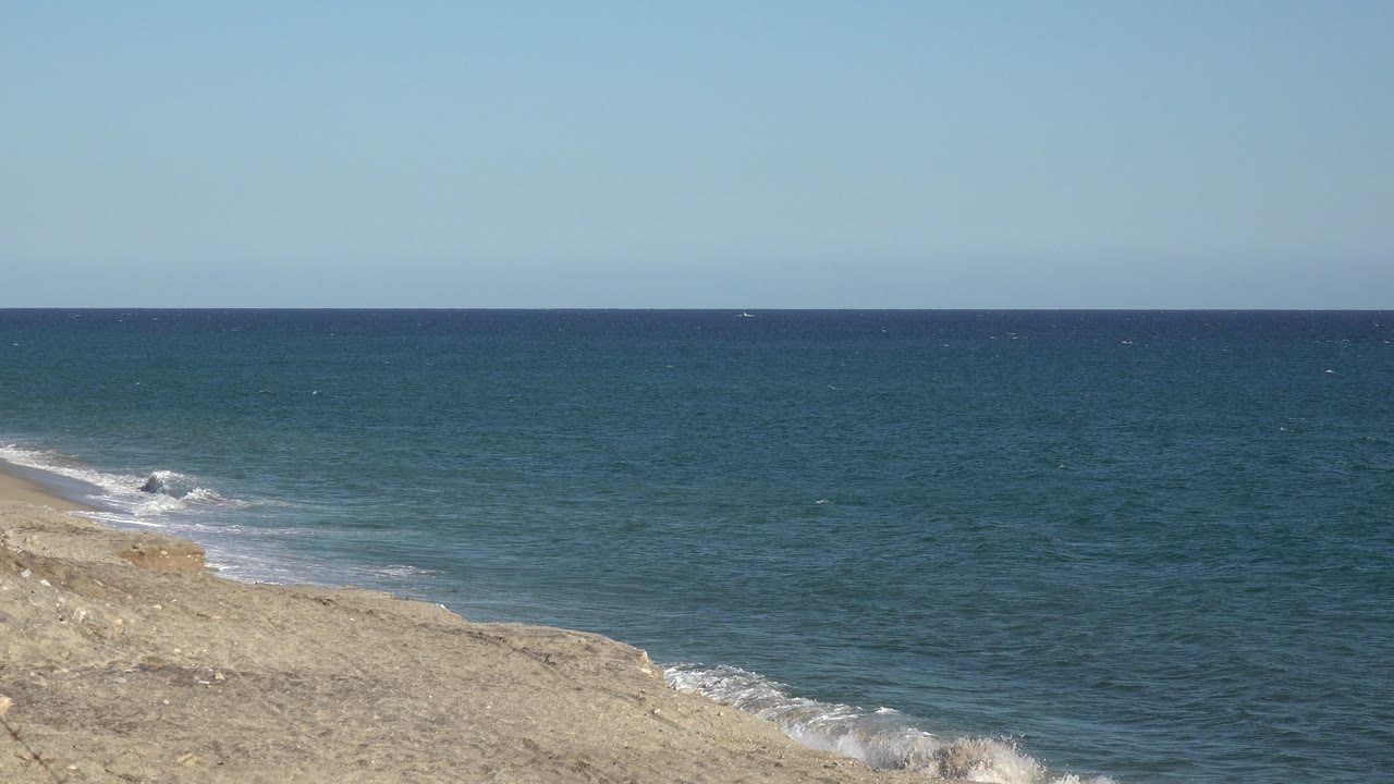 Blue ocean water rolls toward the tan shoreline in Grand Junction Colorado as a small white wave breaks near the left edge, with no visible people.