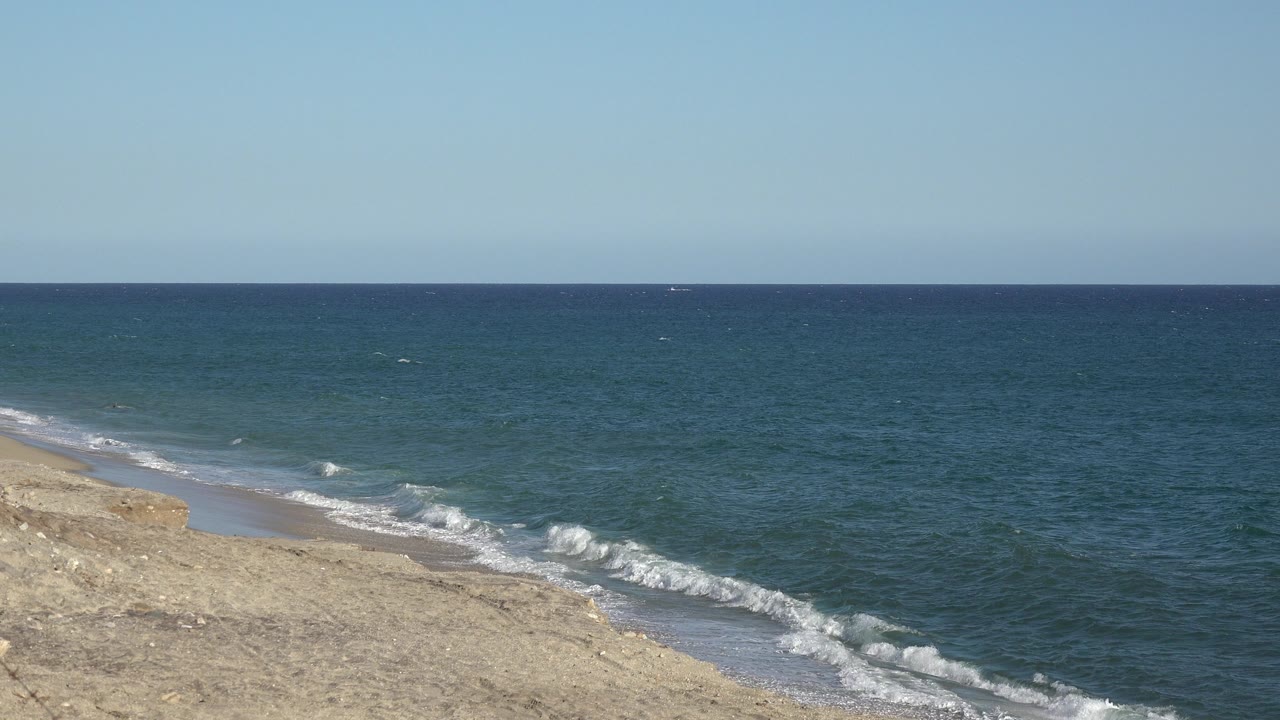 Opening frame of the Grand Junction, Colorado clip, showing a sandy beach at lower left, small surf lines, blue-green open water, a clear horizon, and no visible people.