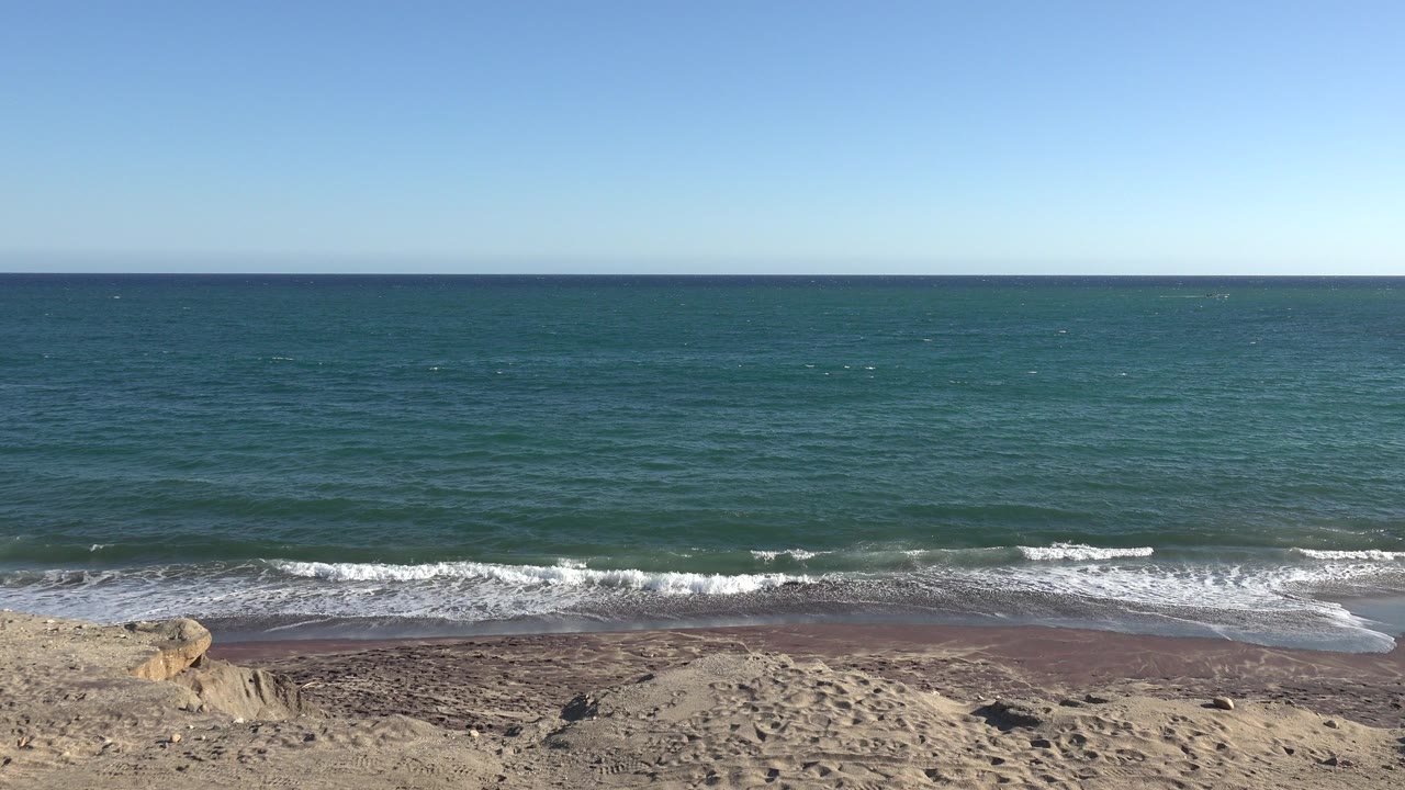 Wide shoreline with blue water, clear sky, sand, rocks, and low waves.