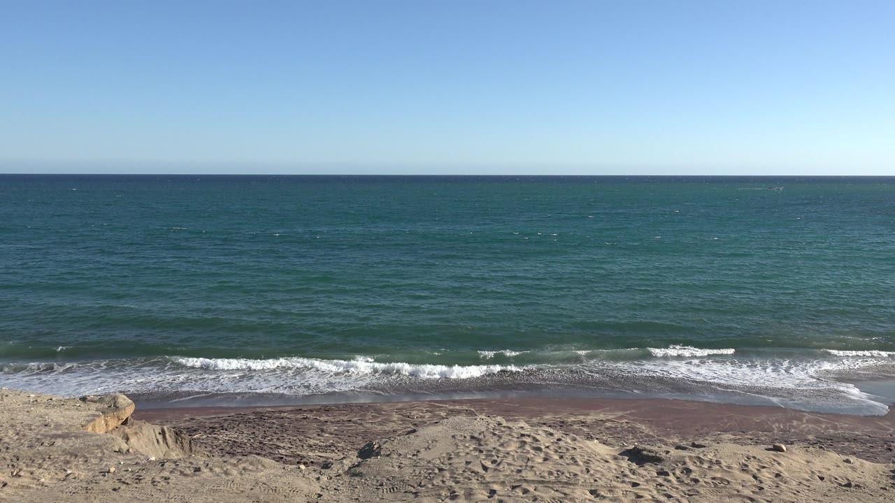 At 0:01, the Grand Junction, Colorado frame holds the level horizon while a small white wave line spreads across the dark beach, with pale sand and a rock outcrop below and no visible people.