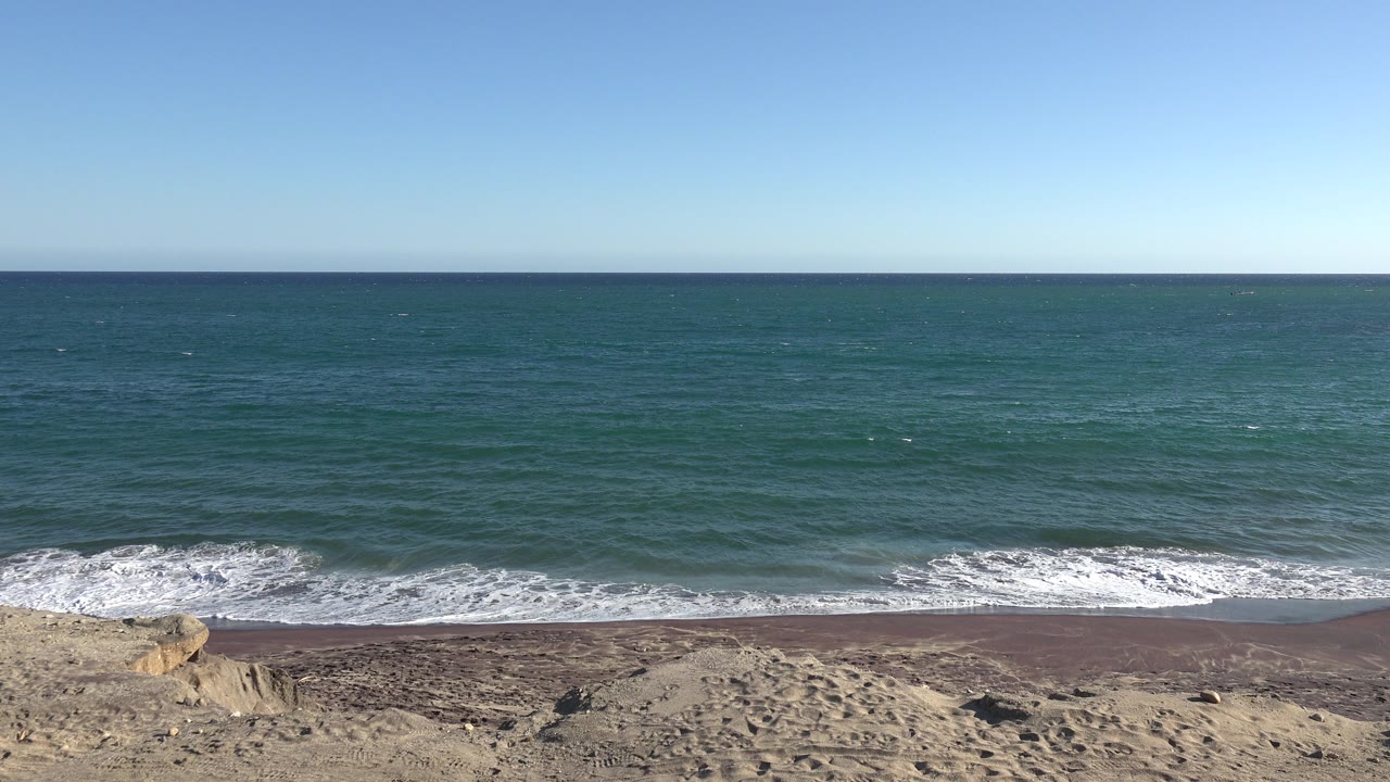 Wide shoreline with rocks, sand, blue water, and clear sky.