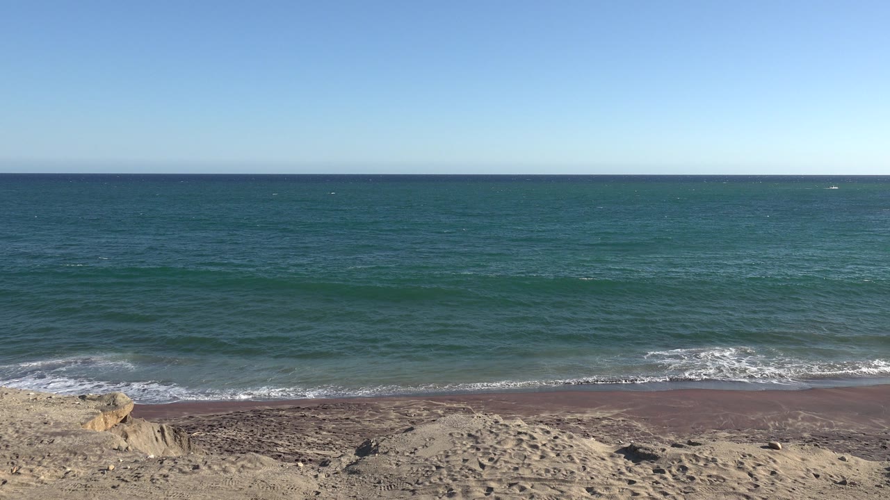 Wide beach view with blue ocean, small waves, sand, and clear sky.