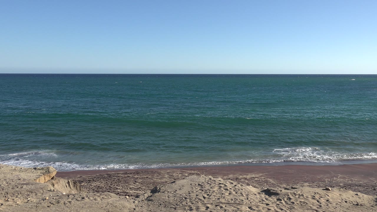 At 0:01, low waves spread across the midground while the sandy foreground and open blue sky remain unchanged, with no visible people.