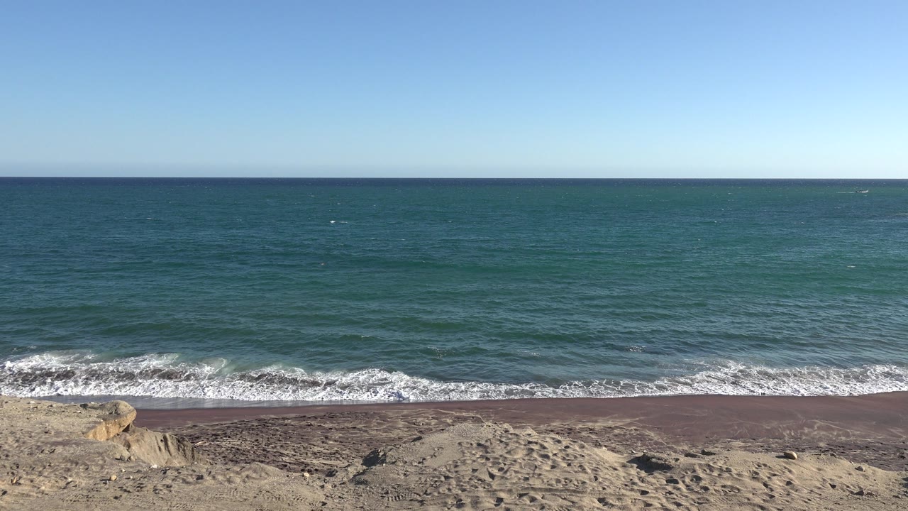 Wide shoreline with low waves, sand, rocks, and blue sky.