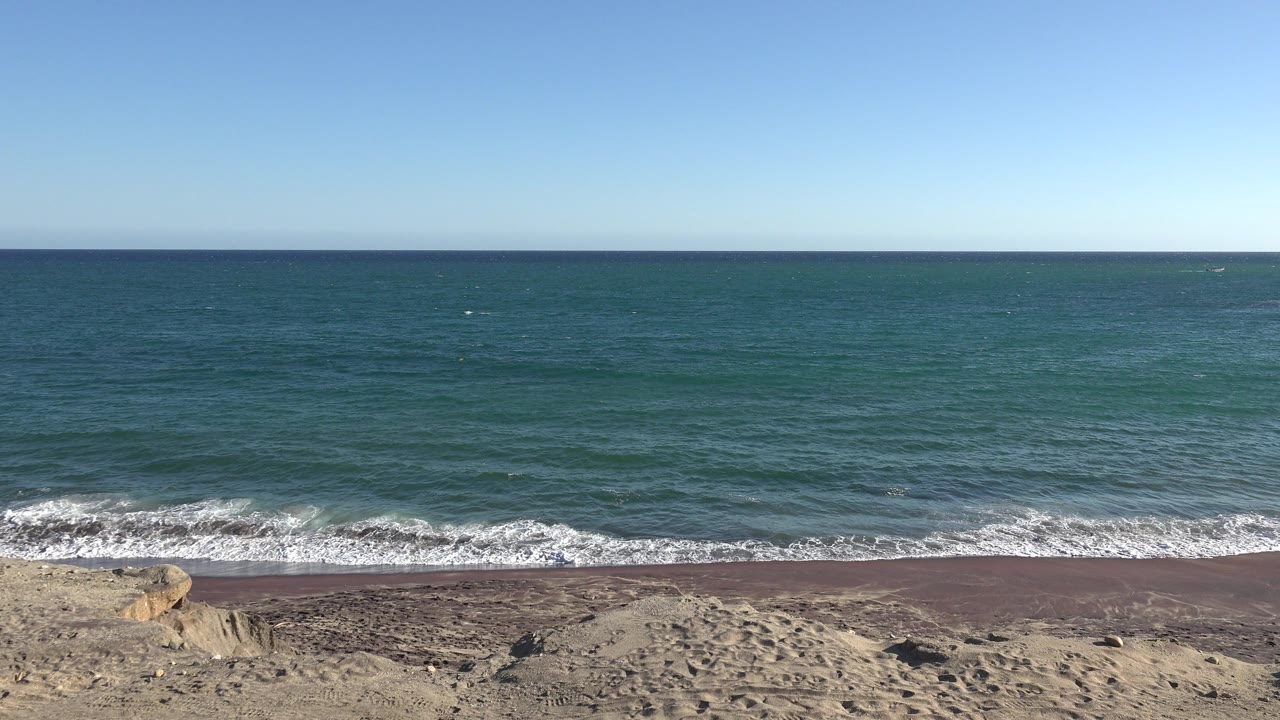 At 0:01, the Grand Junction, Colorado shoreline view shows a thin white wave line spreading along the sand and rocks, with no visible people.