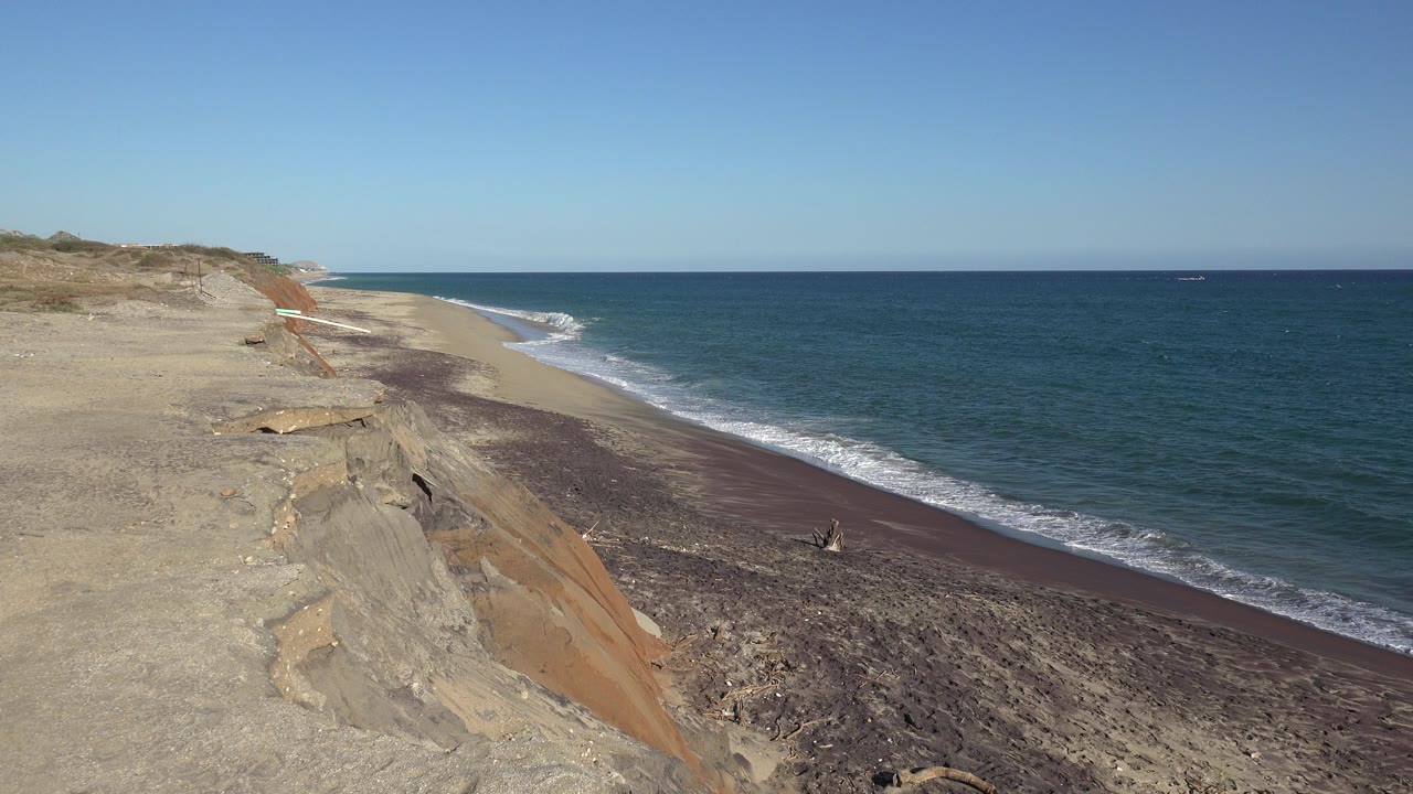 Rocky shoreline below sandy bluffs with blue ocean and sky.