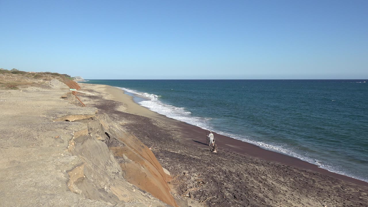 Distant man walking on a narrow beach below sandy bluffs and blue water.