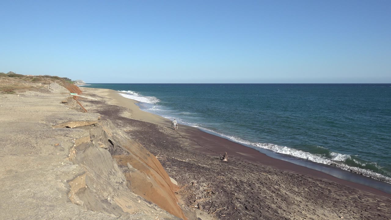 Opening frame of the Grand Junction, Colorado clip, looking from a rocky bluff toward a narrow beach, blue ocean, white surf, and a small distant person near the waterline.
