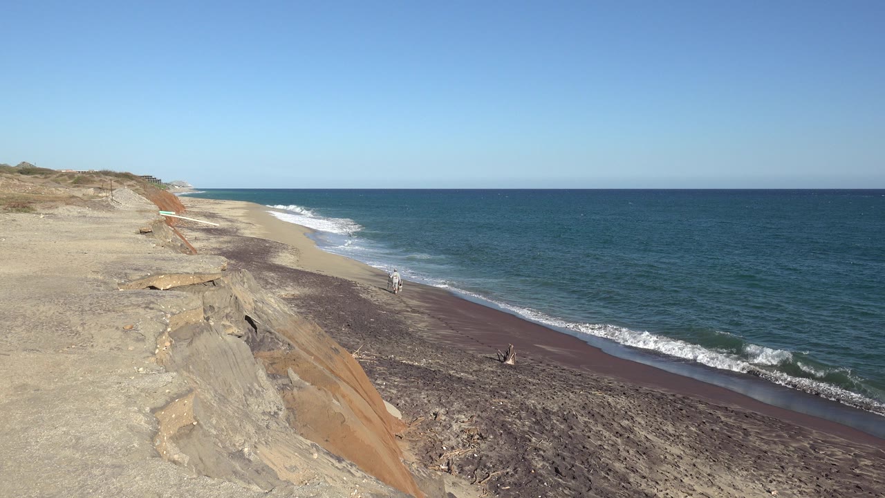 At 0:01, the Grand Junction, Colorado shoreline view holds on the bluff at left, a darker pebble strip along the beach, foamy waves, and tiny people near the surf.
