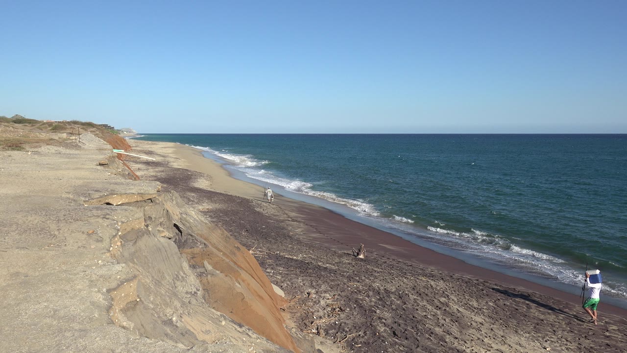 At 0:00, an elevated Grand Junction, Colorado beach view shows eroded sand at left, blue water at right, and small people walking along the sunlit shoreline.