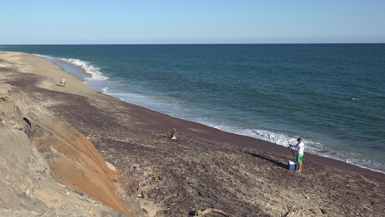 Wide elevated view in Grand Junction Colorado collection showing a person near a blue cooler on the lower right shoreline as waves roll onto dark sand beneath a tan bluff.