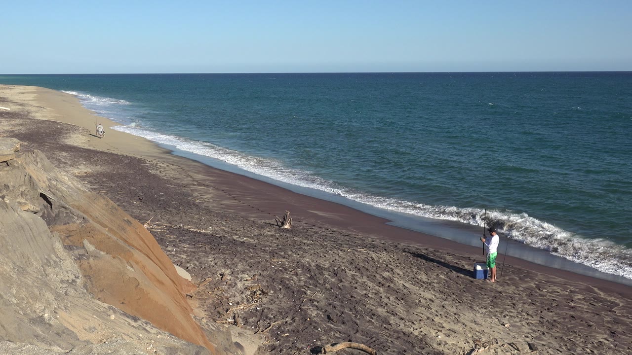 Person standing on a beach below a bluff as waves reach the shore.