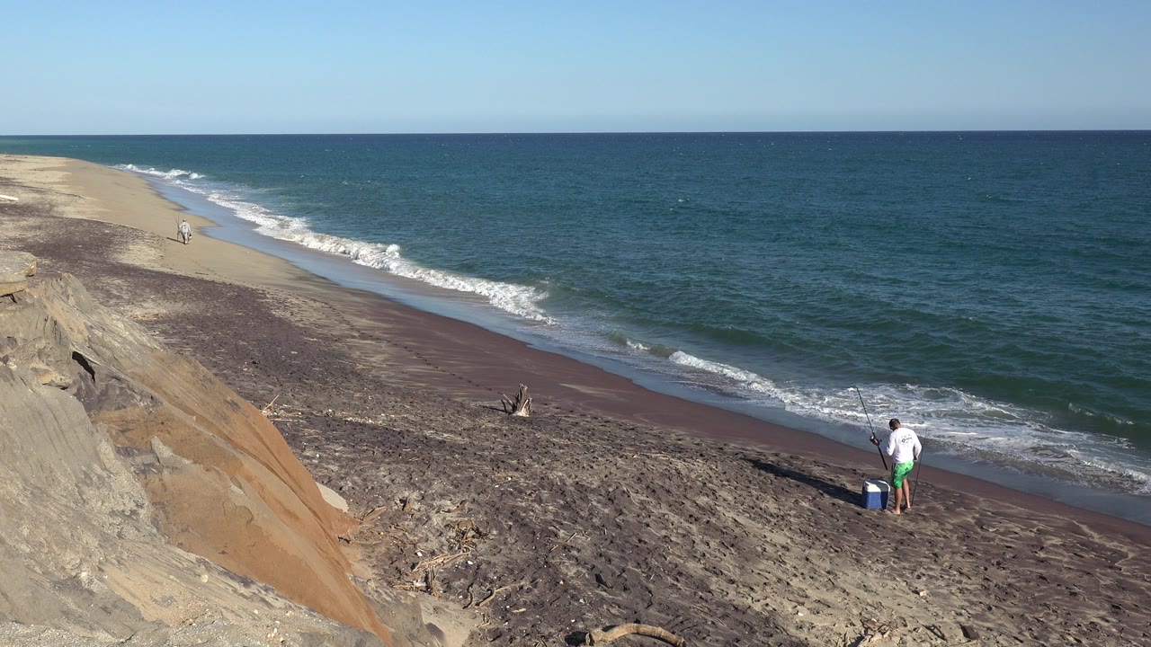 Wide shoreline view with a distant man fishing beside a blue bucket.