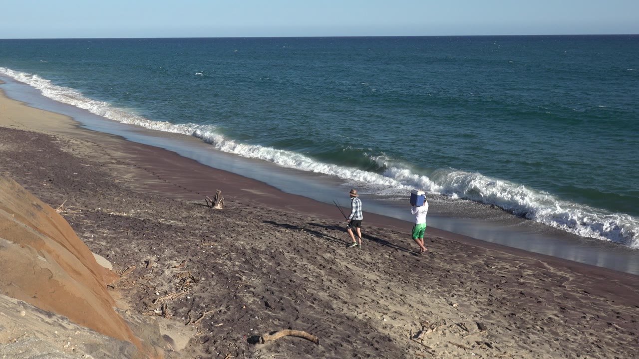 Two people on a sloped sandy beach beside waves and blue water.