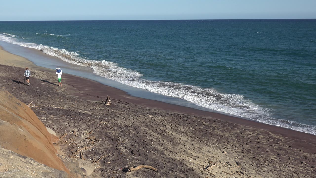 At 0:00, two people walk along the Grand Junction, Colorado shoreline below a sandy bluff as small waves break on dark wet sand.