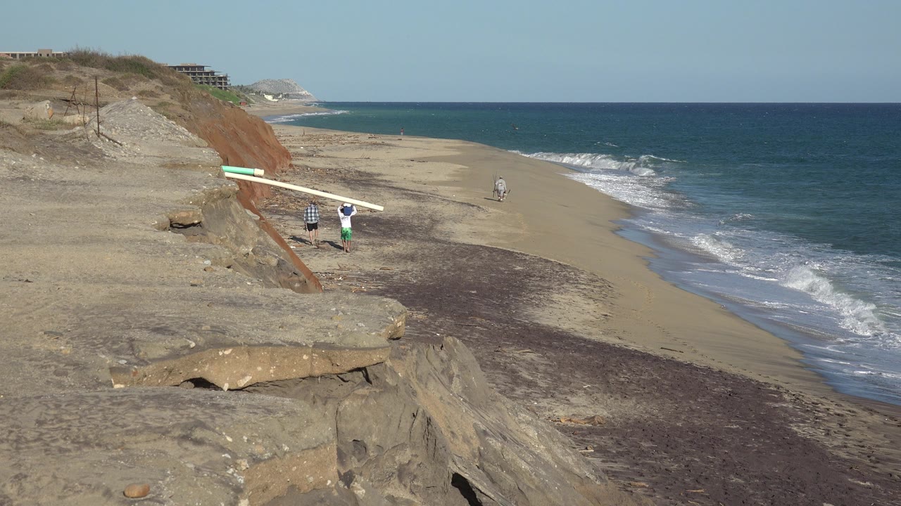 At 0:01, the Grand Junction, Colorado shoreline remains framed from above, with the two people and long board slightly farther along the beach beneath the sandy bluff.