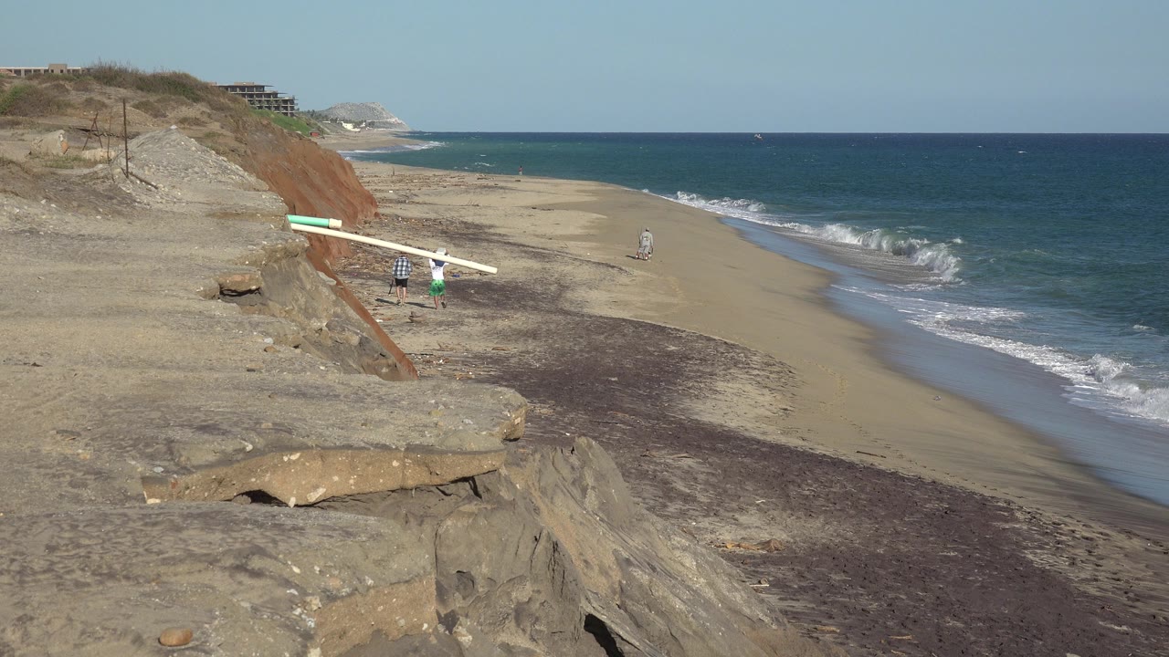 Opening frame of a Grand Junction Colorado 2017 clip showing tan eroded bluffs, a broad sandy beach, blue ocean, and two small distant people near long white boards.