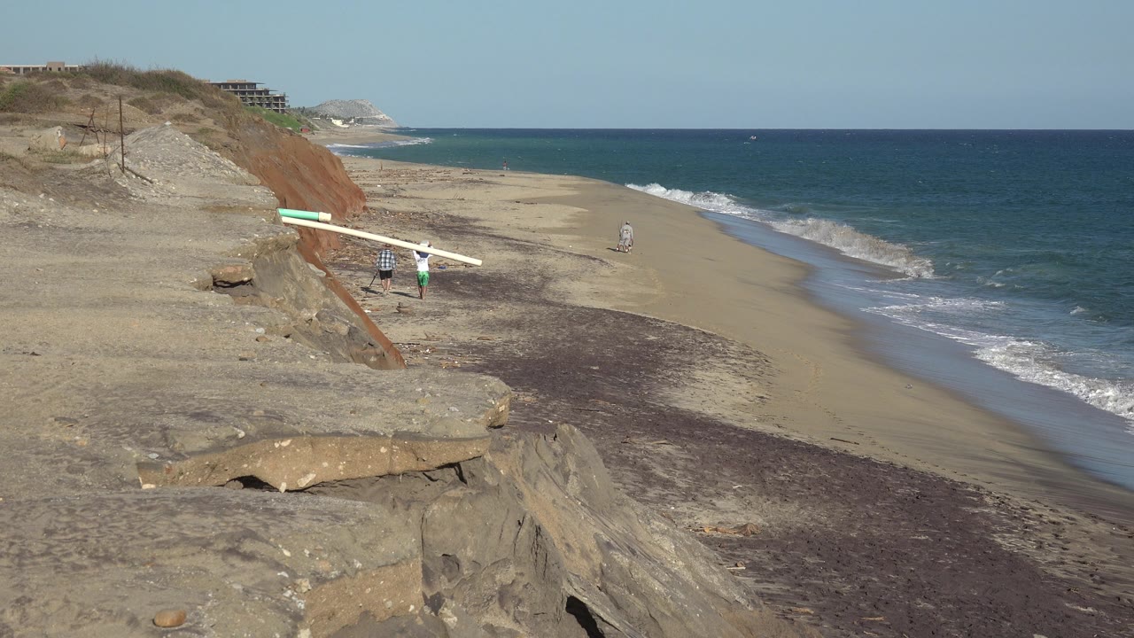 Wide shoreline view with blue water, tan bluff, waves, and two small beach figures