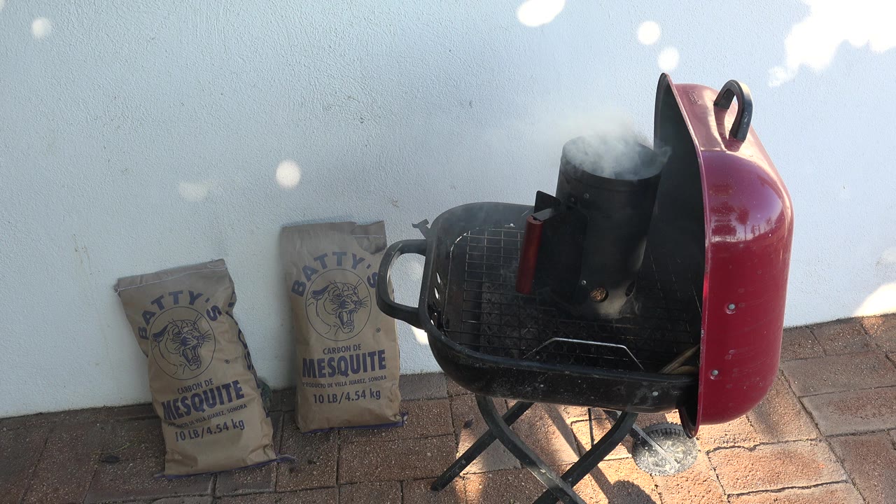 A red kettle charcoal grill in Grand Junction, Colorado holds a black chimney starter giving off smoke beside two mesquite charcoal bags on a brick patio, with no visible people.