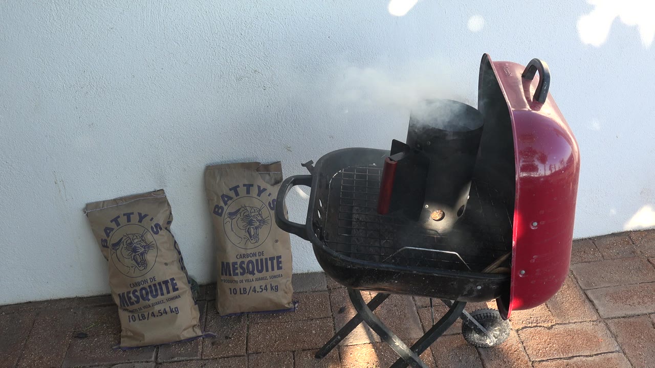 Red charcoal grill with smoking chimney starter beside mesquite charcoal bags on a patio.