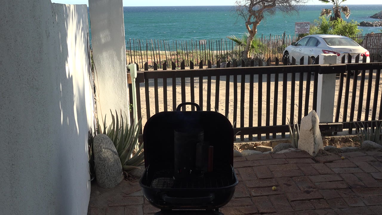 Black barbecue grill on a shaded patio with a white car and water beyond a fence.