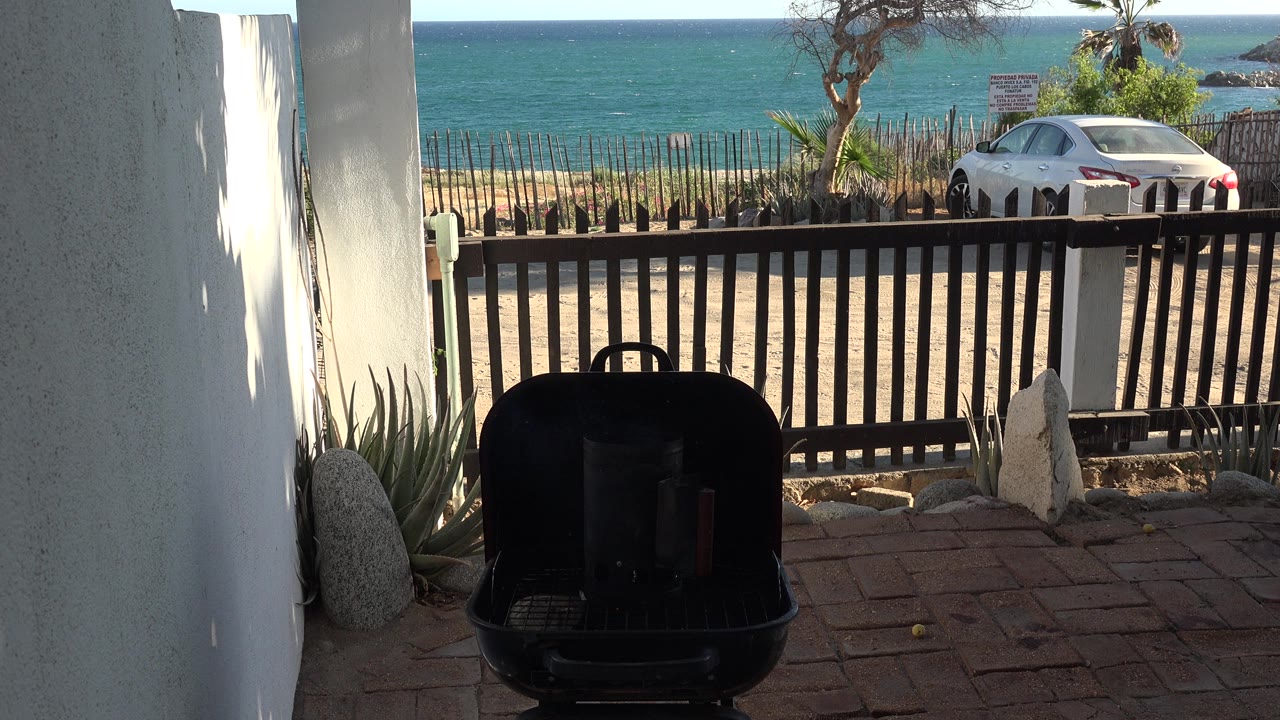 Shaded patio grill facing a fence, parked car, beach, and ocean.