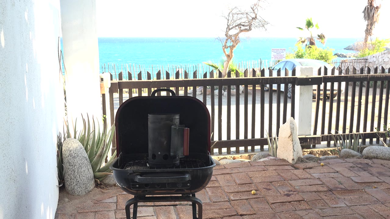At 0:00, an open black grill stands on a brick patio in Grand Junction, Colorado, with white walls, a dark fence, trees, bright turquoise water, and no visible people.
