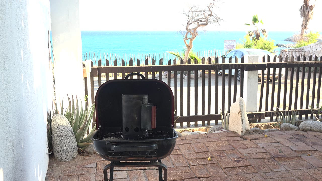 Black grill on patio with white walls, fence, trees, and bright water behind it.
