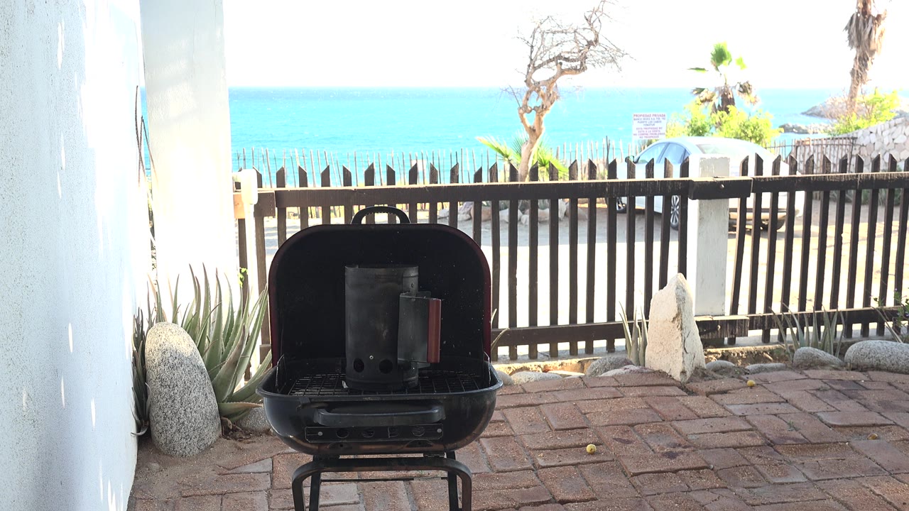 Open black grill on a brick patio with a fence and blue ocean beyond.