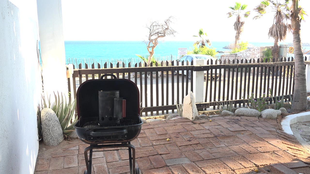 At 0:01, the Grand Junction Colorado frame keeps the black grill centered lower in view, with brick paving, fence rails, palm trunks, dry landscaping, and blue water in harsh daylight, with no visible people.