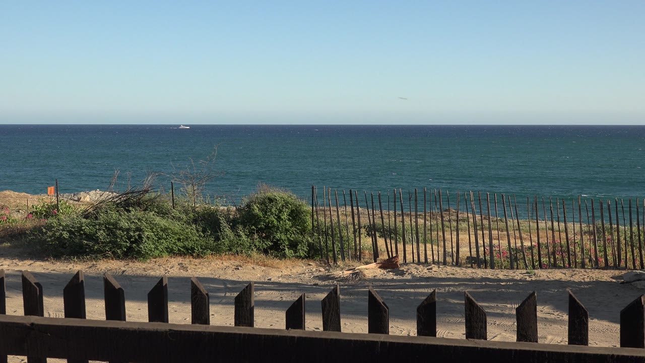 Open water with small distant boat, sandy bank, brush, fence, and dark railing.