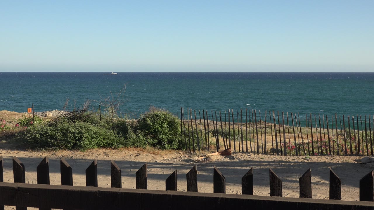 Grand Junction Colorado 2017 frame with the horizon stretching across blue water, low beach shrubs, thin fence posts, dark foreground rail, and no visible people.