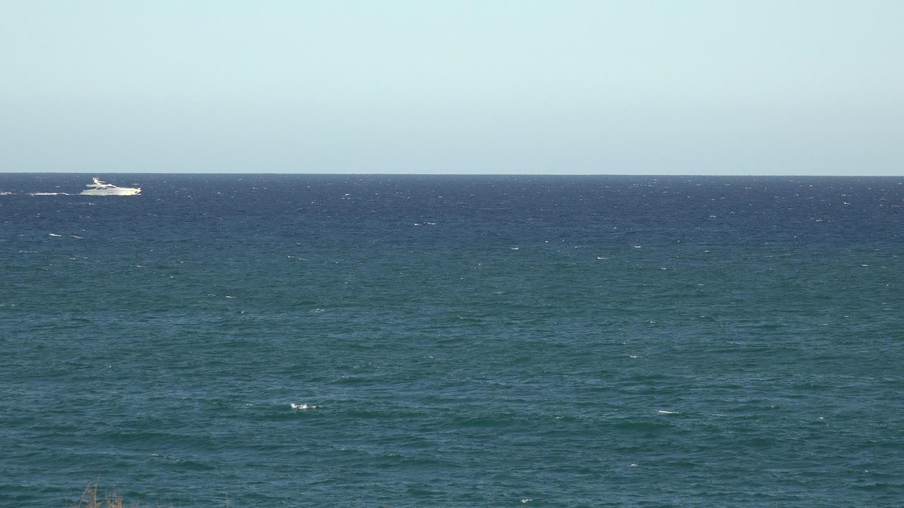 Open blue water under a clear pale sky, with a small white boat closer to the center horizon and no visible people.