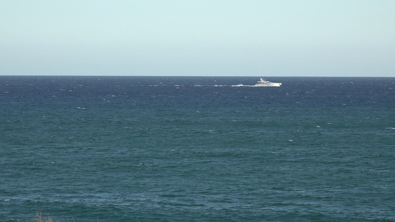 The distant white boat sits closer to the center of the horizon line, with textured teal water below, pale blue sky above, and no visible people.