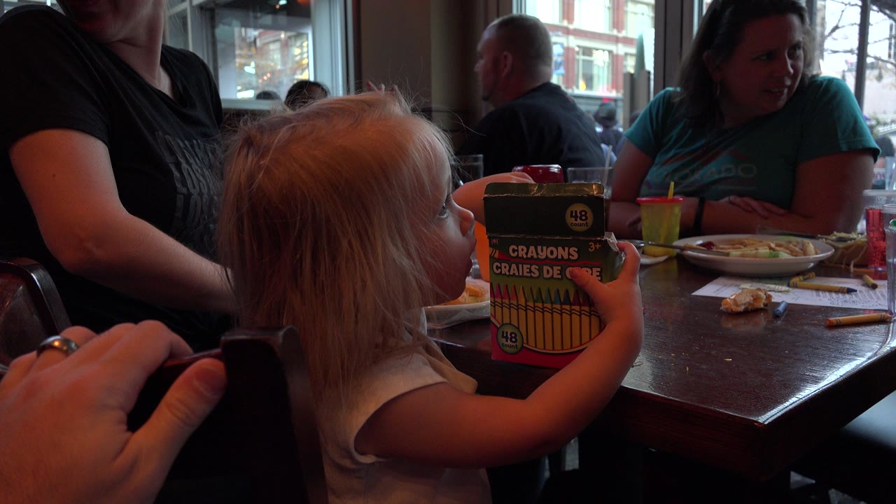 In Denver, Colorado, a young child sits at a restaurant or mall table holding a crayon box while adults sit nearby in window light.