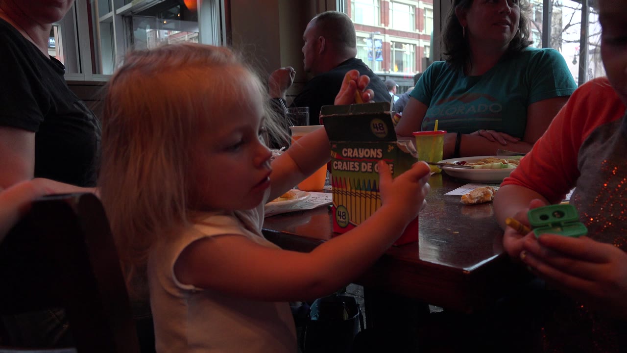 A young blonde girl sits at a restaurant table in Denver, Colorado, holding a crayon box while adults and window light fill the background.