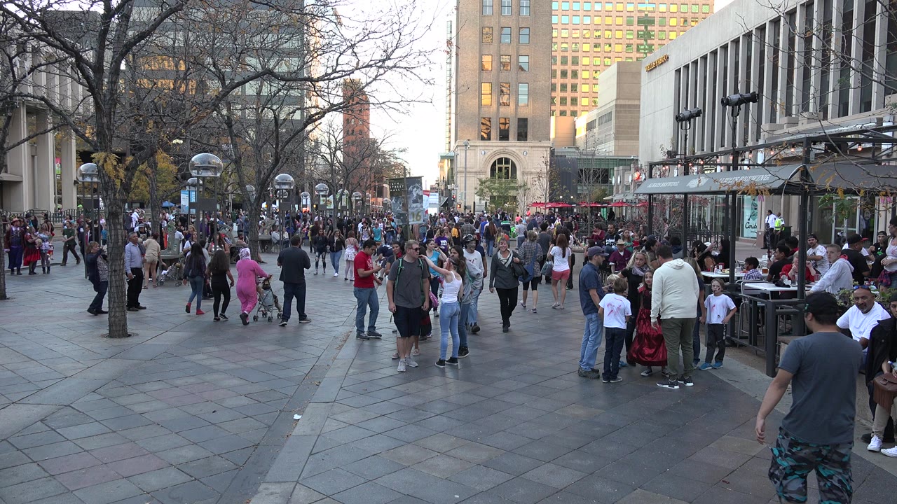 Wide view of a Denver paved plaza with crowds of adults and children walking between leafless trees, lamp posts, patio tables, and tall buildings.