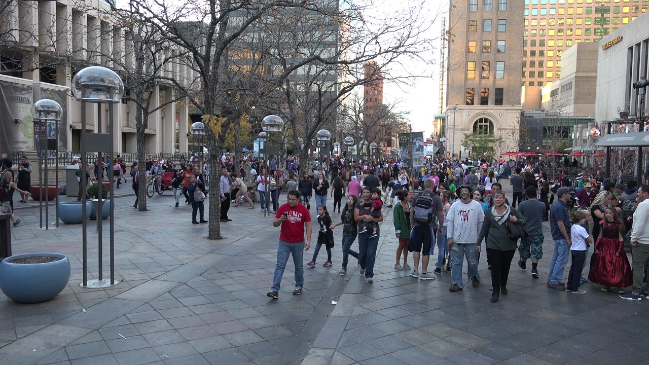 Crowd walking through a paved city plaza with tall buildings and globe lamps.