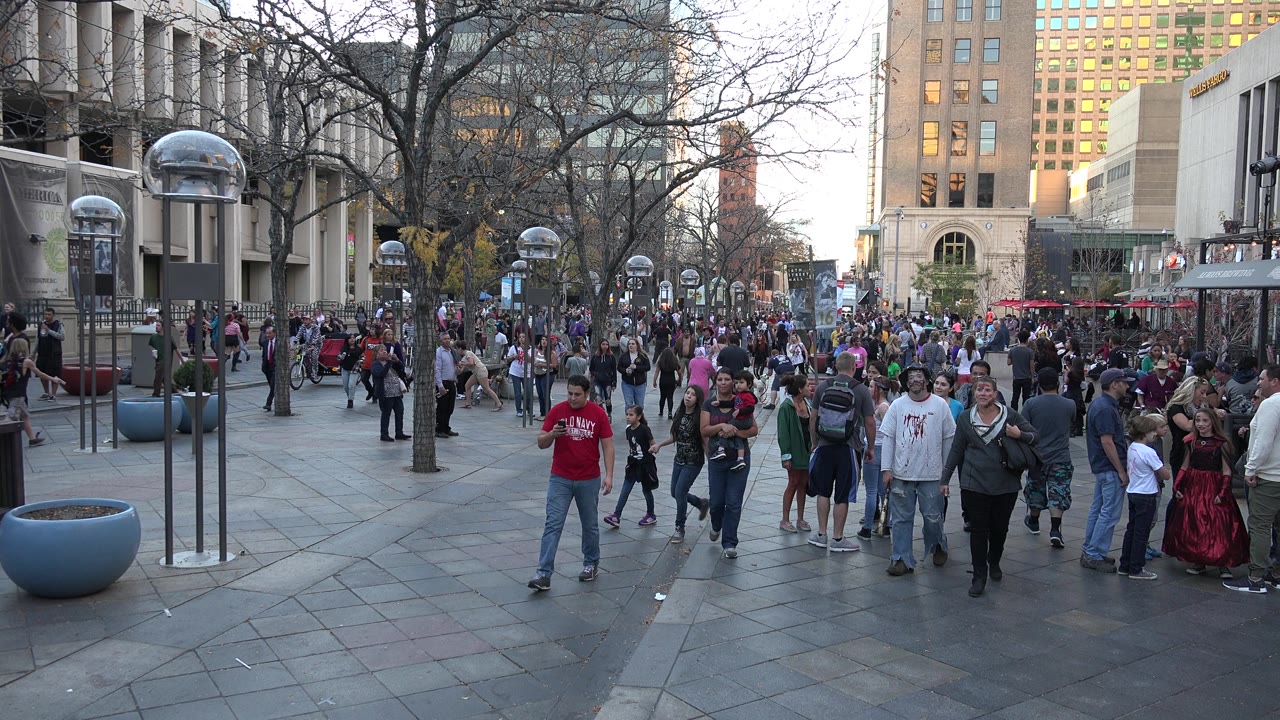 People continue walking through the Denver plaza, with a red-shirted man in the foreground, families nearby, a dense crowd behind them, and museum-style signage along the left building.