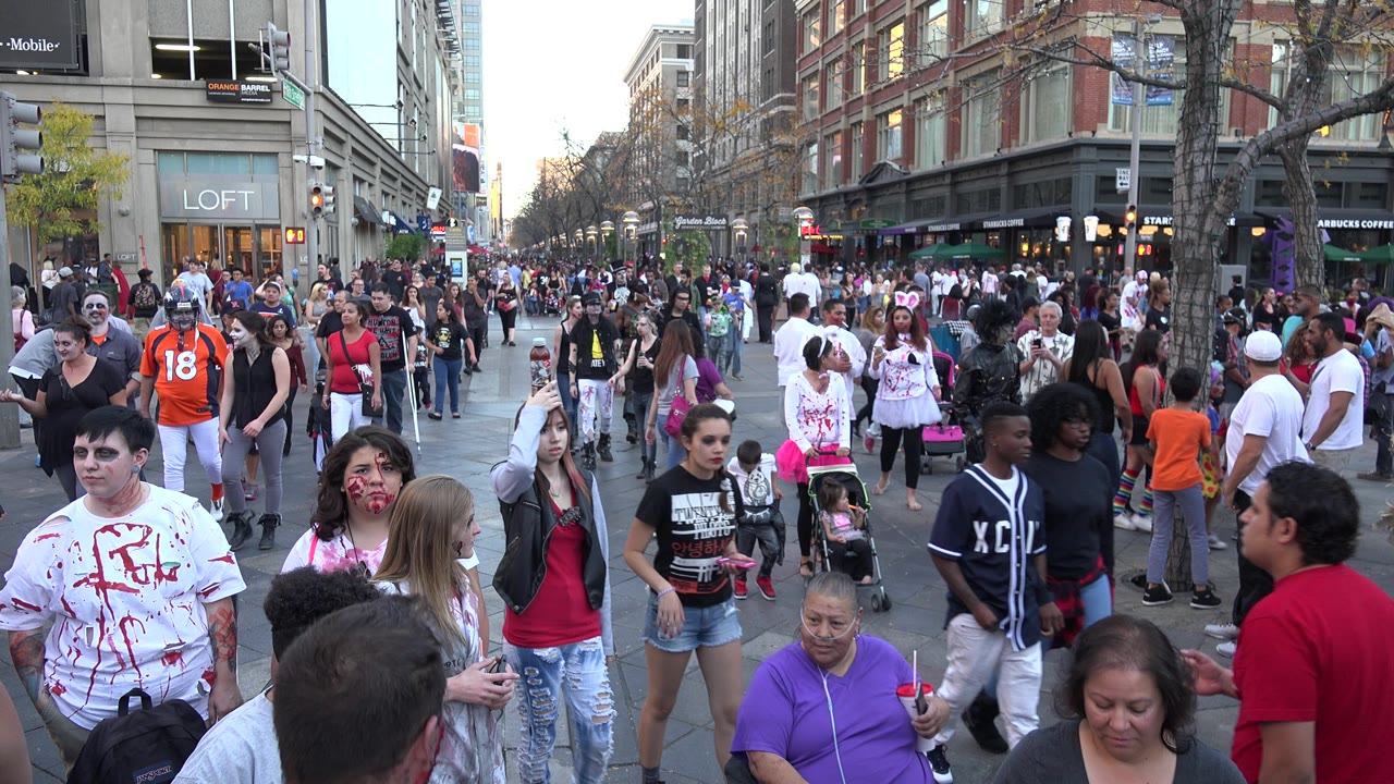 Denver street view shows a dense pedestrian crowd with a man in an orange jersey, people in zombie-style makeup, storefronts, bare trees, and shop signage along both sides.